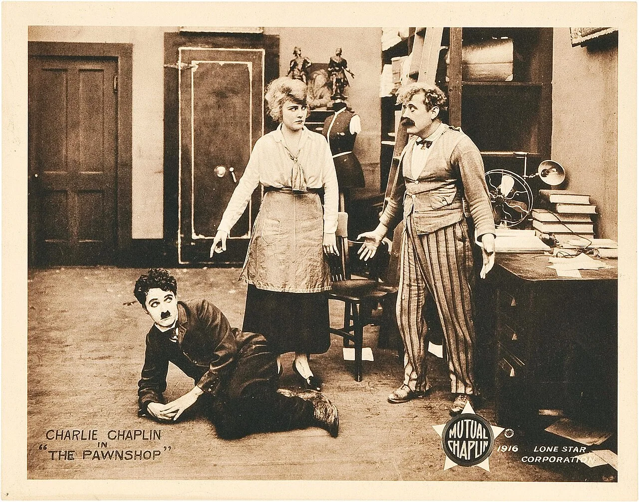 بطاقة عرض لفيلم The Pawnshop: تشابلن على الأرض وشخصان داخل الدكان.