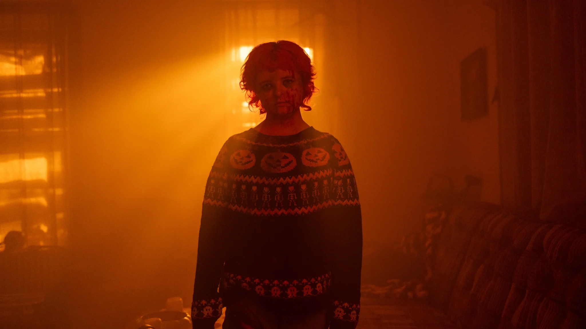 بامبكن بآثار دم على وجهها تقف وسط ضباب وإضاءة برتقالية قوية داخل المنزل