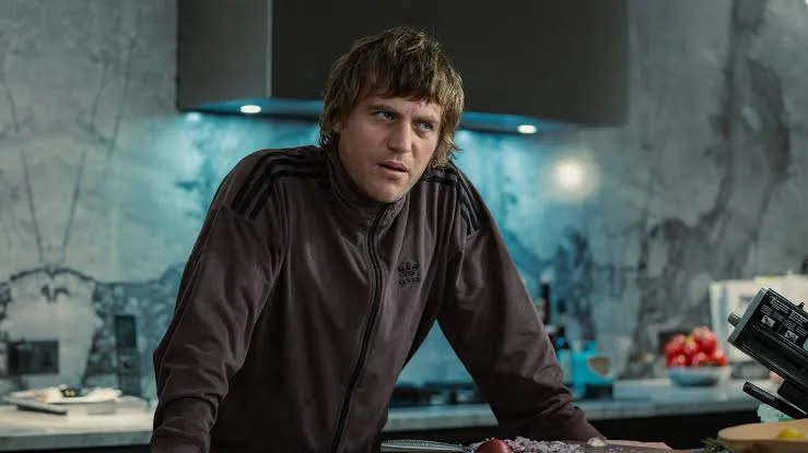 إيدي يميل إلى الأمام بجوار منضدة مطبخ مرتديًا قميصًا داكنًا